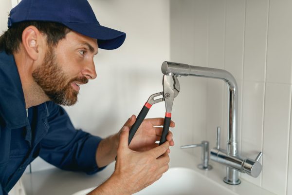 Professional plumber in blue uniform fixing broken water tap in kitchen. High quality photo